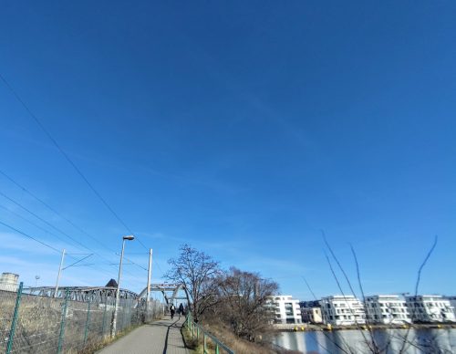 EIn Brückenweg über ein Gewässer, viel blauer Himmel, in der Ferne ein paar Wohnhäuser.