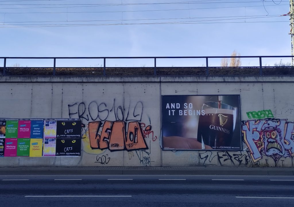 Wand mit Graffiti und Werbungen, eine davon groß, ein Guiness-Bierglas und der Spruch "And so it Begins.."