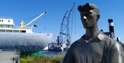Hafenimpression, im Vordergrund die Statue eines Arbeiters, im Hintergrund ein Kran und ein Schiff