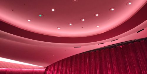 Blick an die Decke eines Kinosaals. Alles ist in Rottönen gehalten, einzelne Lichtstrahler sind in die Decke eingelassen, eom Vorhang aus rotem Samt vor der Kinoleinwand ist noch halb im Bild.