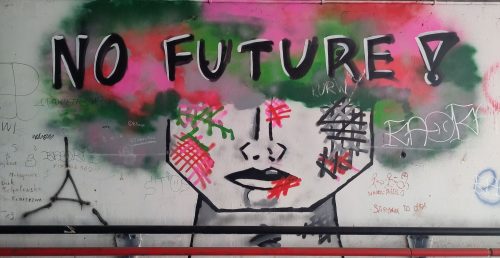 Bild eines Grafitto, auf dem ein zur Hälfte von dunkel-bunten Haaren verdecktes Gesicht abgebildet ist. Quer darüber steht "No Future!"