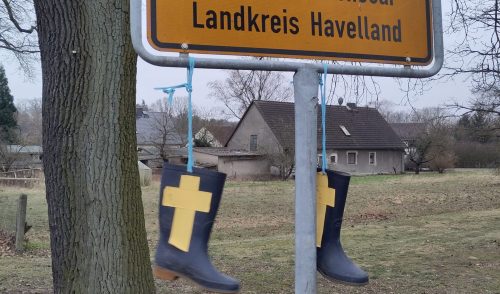 An einem Ortsschild von dem nur die Zeile "Landkreis Havelland" sichtbar ist, hängen zwei Gummistiefel, auf die gelbe Kreuze geklebt sind.