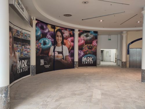 Ausschnitt eines runden, leeren Raums, mit einer geöffneten Tür am fernen Ende, die in einen ebenfalls leeren Gang führt. Im Bogen zu linker Hand generische Werbung den Verkauf von Backwaren (es gibt kein entsprechendes Geschäft im Center), ganz rechts die geschlossene Tür zu einem Fahrstuhl. Der Boden ist entfließt und aufgeraut.